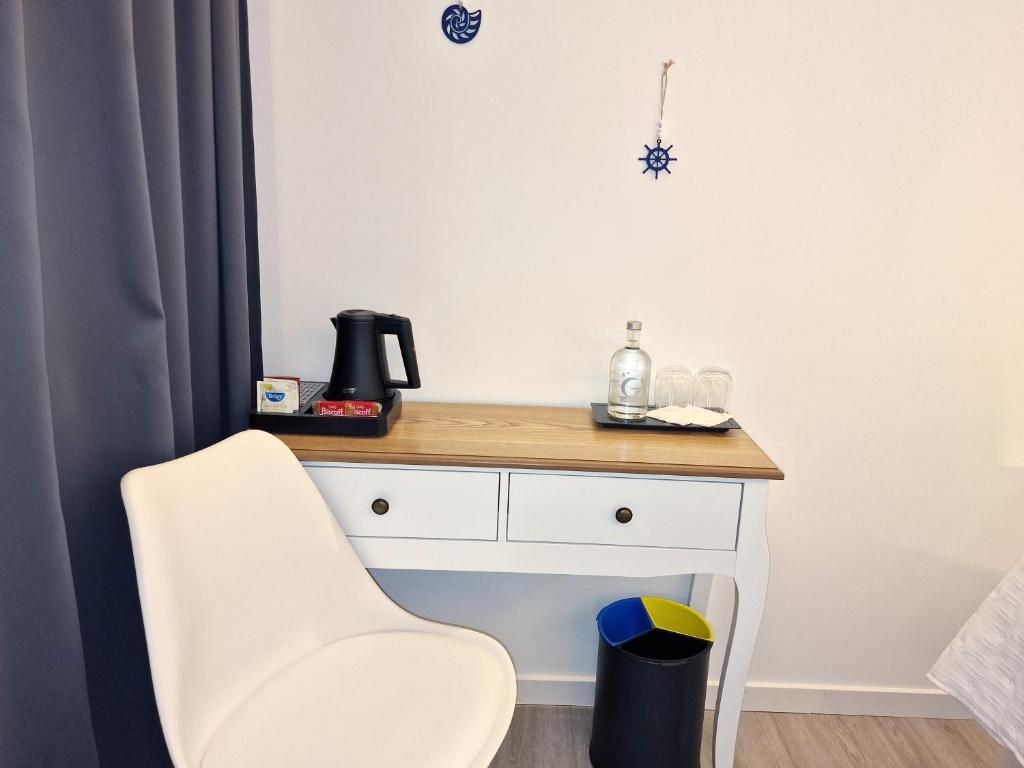 A hotel room with a white chair, a wooden desk, a coffee maker, a bottle, and glasses.