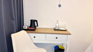 A hotel room with a white chair, a wooden desk, a coffee maker, a bottle, and glasses.