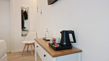 Una pequeña cocina americana con cafetera, hervidor de agua y una bandeja con tazas y un vaso. Hay una silla blanca y un sofá blanco en la habitación.