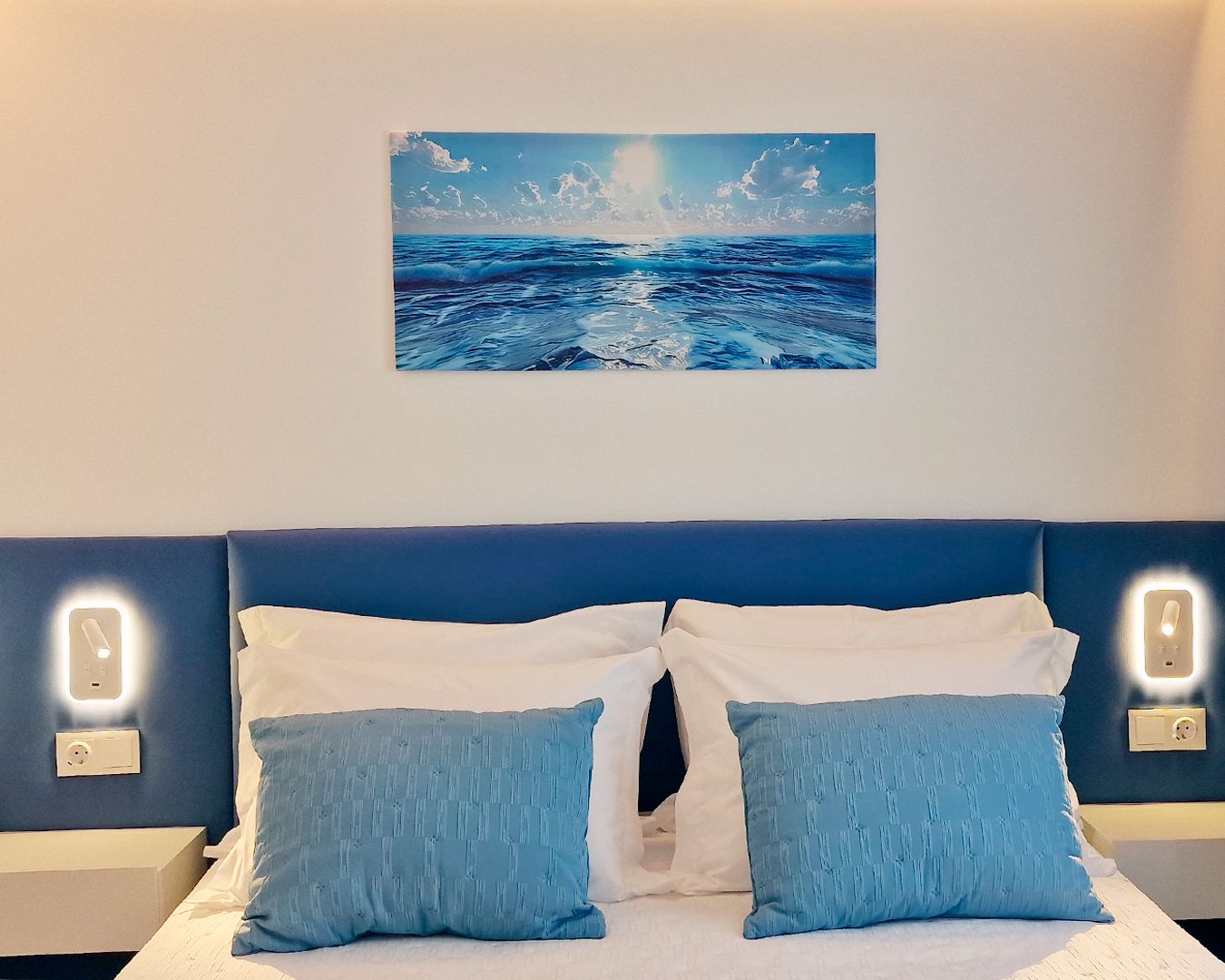 Un dormitorio con cama, dos almohadas, un cuadro colgado en la pared que representa el océano y lámparas de noche.