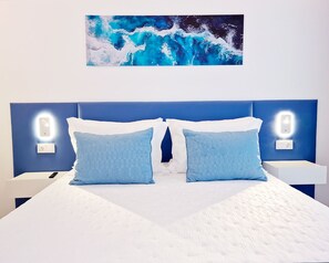 A bed with a white mattress, two blue pillows, and a blue headboard. There are two bedside tables with illuminated switches. - Golfinho Azul (Odemira)