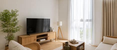 Un salon moderne avec un téléviseur à écran plat, une table basse en bois, un lampadaire, une grande fenêtre avec des rideaux légers et une plante en bambou.