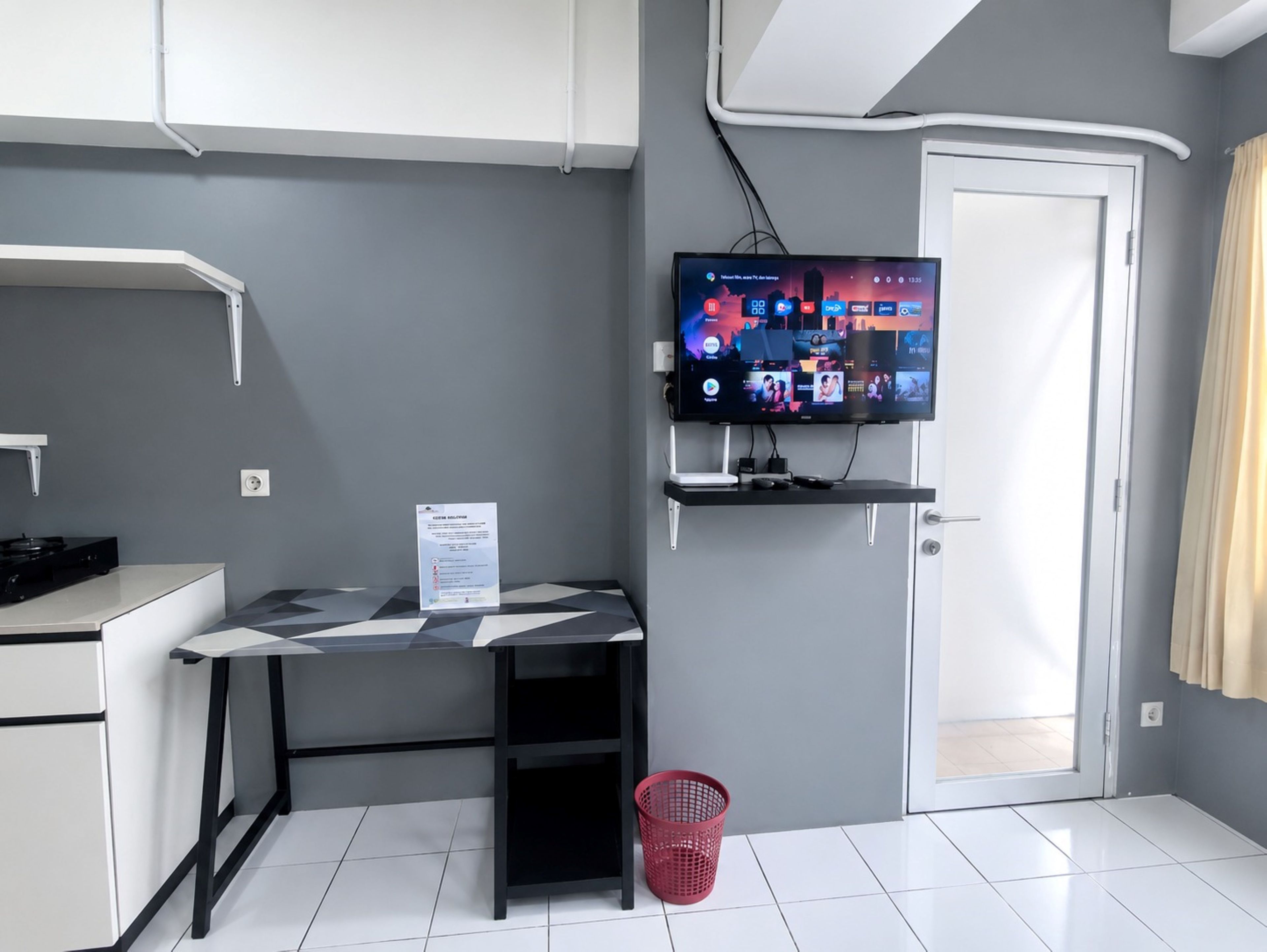 A modern room with a flat-screen TV mounted on the wall, a desk with a chair, a small trash bin, and a door.