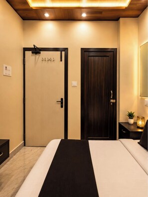 A hotel room with a bed, a wooden door, a nightstand with a lamp, and a wall-mounted mirror. - Divyam Grand (Varanasi)