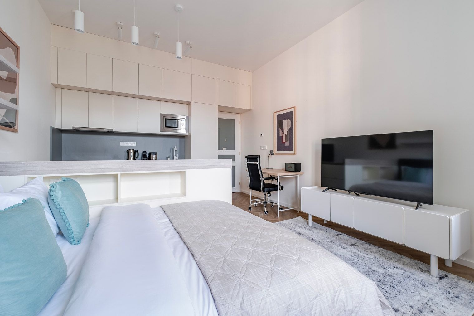 A modern hotel room with a bed, a desk, a television, and a kitchenette.