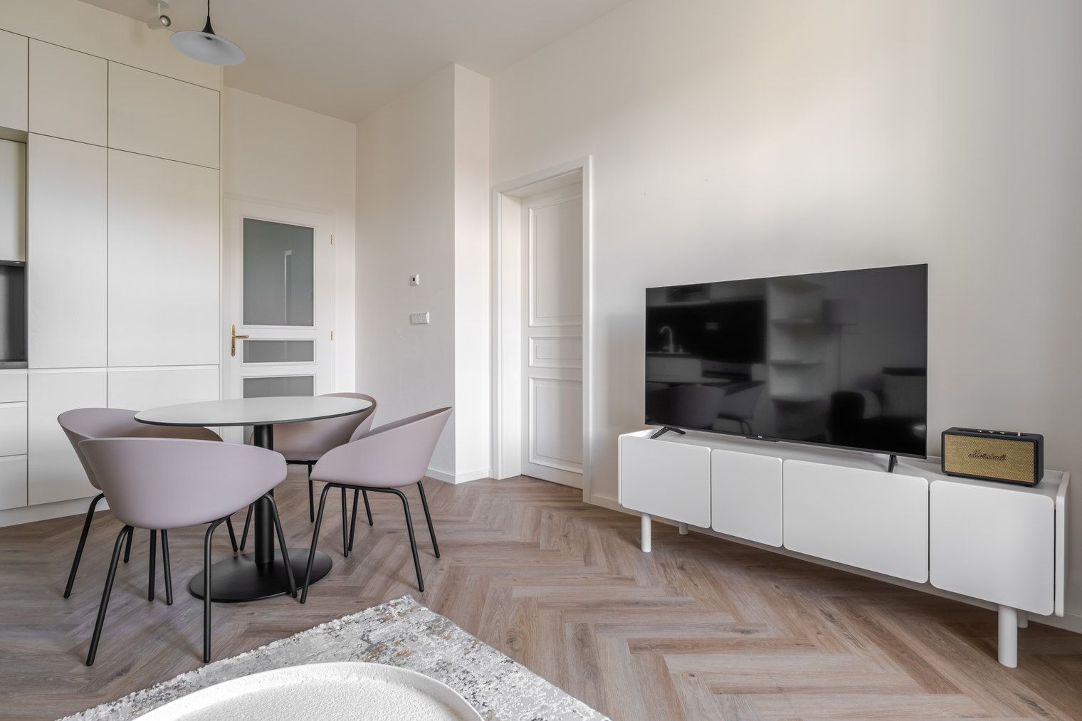 A modern living room with a flat-screen TV on a white cabinet, a dining area with a table and chairs, and a herringbone-patterned wooden floor.