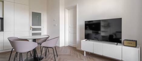 A modern living room with a flat-screen TV on a white cabinet, a dining area with a table and chairs, and a herringbone-patterned wooden floor.