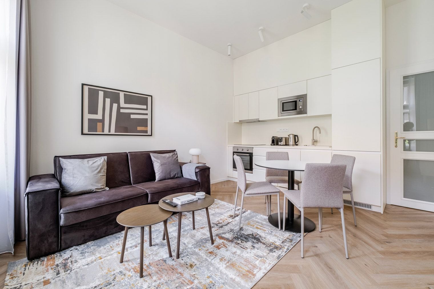 A modern living room with a sofa, coffee table, and chairs, a kitchen with white cabinets and appliances, and a framed artwork on the wall.