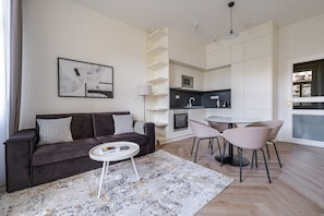 A modern living room with a dark brown sofa, a small white coffee table, and a grey patterned throw pillow. There is a kitchen area with white cabinets, a black countertop, and a round dining table with four chairs. - Vinohrady Residence by Blueground (Prague)