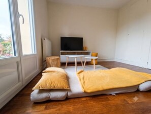 Een kamer met een groot raam, een flatscreen televisie, een klein rond tafeltje en een bank met kussens.
