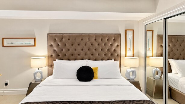 A hotel room with a large bed, a tufted headboard, two bedside lamps, a mirror, and a patterned bedspread.