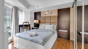 A modern hotel room with a large bed, a desk, a chair, a bedside table, and a window with curtains.