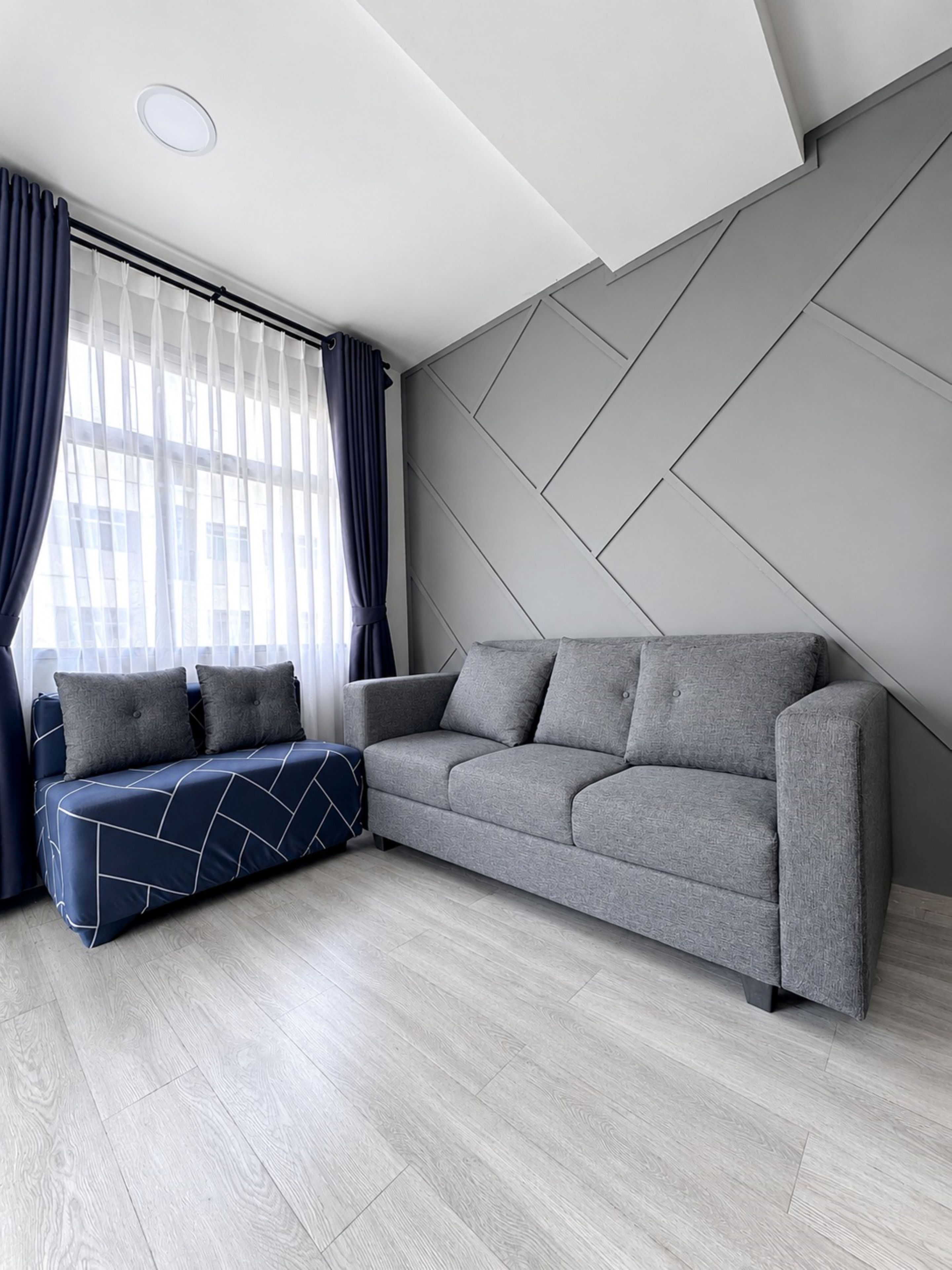A modern living room with a grey sofa, a patterned ottoman, and a geometric wall design.