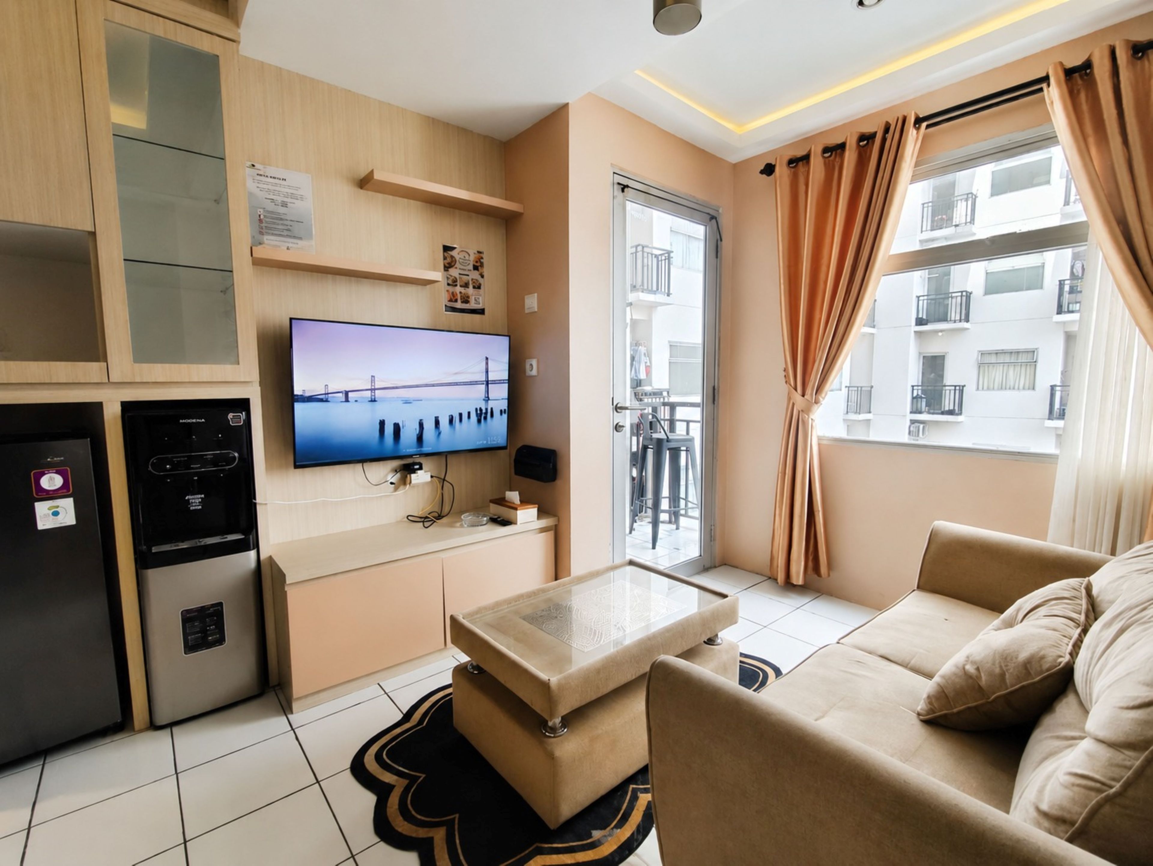 A modern living room with a flat-screen TV, a sofa, a coffee table, and a kitchen area with a refrigerator and a microwave.