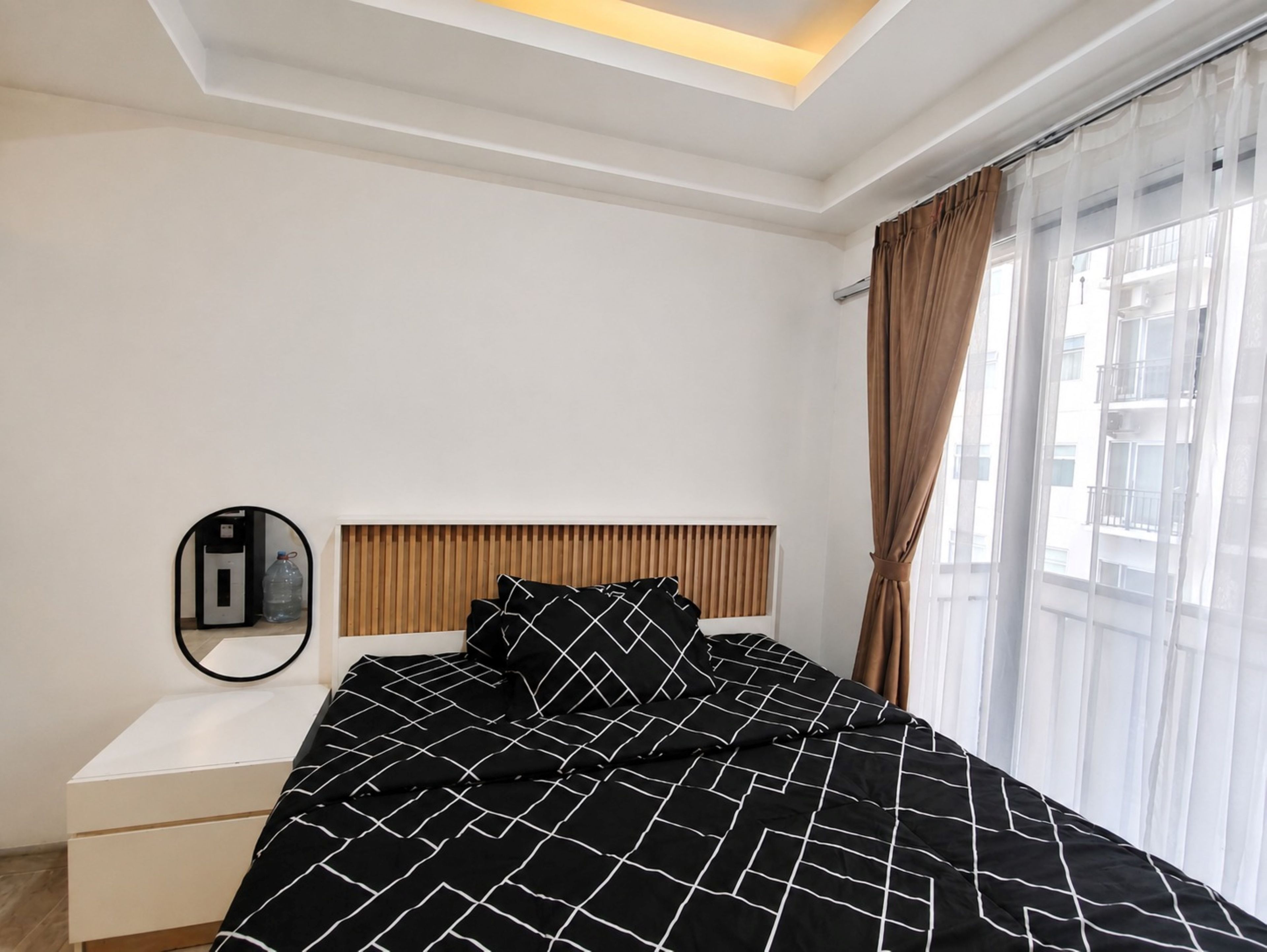 A bedroom with a bed, a bedside table, a mirror, and a window with curtains.