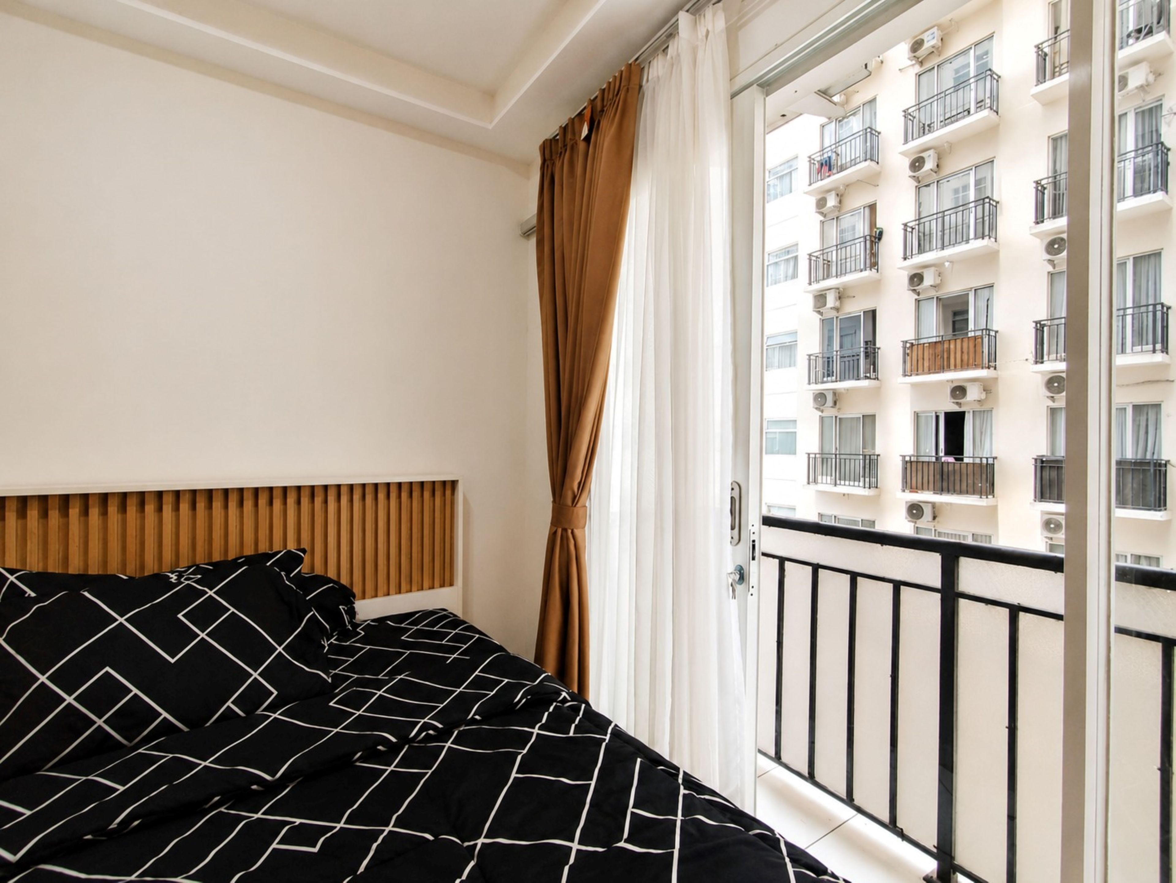 Ein Hotelzimmer mit Bett, Holz-Kopfteil, Fenster mit Vorhängen und Blick auf ein Gebäude mit Balkonen.