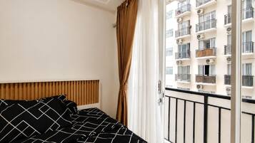Ein Hotelzimmer mit Bett, Holz-Kopfteil, Fenster mit Vorhängen und Blick auf ein Gebäude mit Balkonen.