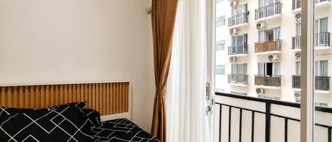 A hotel room with a bed, a wooden headboard, a window with curtains, and a view of a building with balconies.