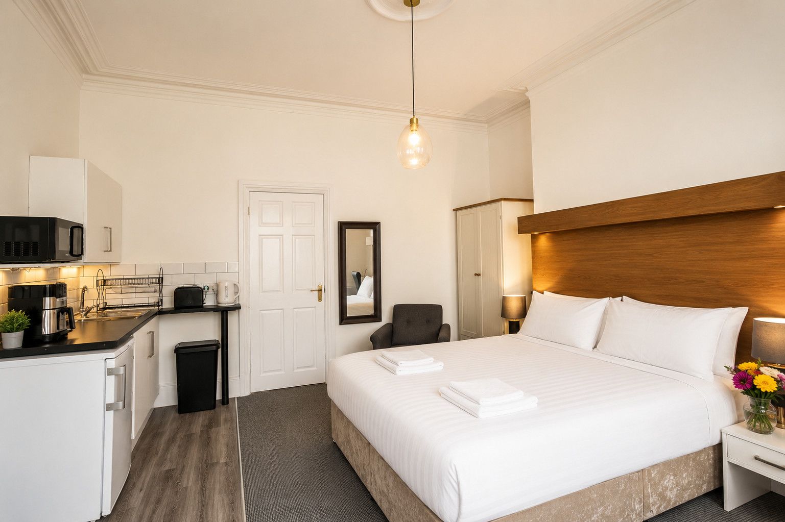 Ein Hotelzimmer mit Bett, Nachttisch, Lampe und einer Küchenzeile mit Mikrowelle und Kaffeemaschine.