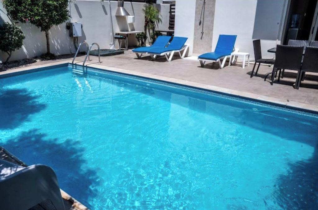 Una piscina con escalera, sillones reclinables azules y una mesa con sillas.