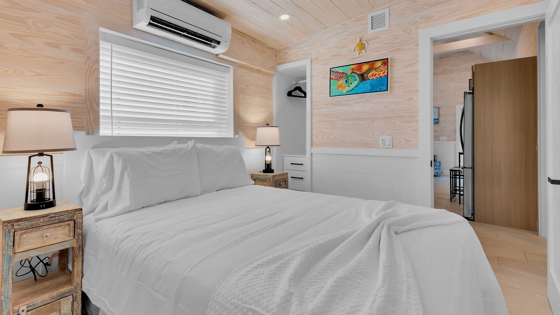 Un dormitorio con cama, lámparas de noche, un equipo de aire acondicionado, un cuadro y una puerta que da a otra habitación.