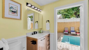 A bathroom with a white sink, wooden cabinet, and a large mirror. There is a view of an outdoor pool area with chairs and a fence. - **SECRET SAND DOLLAR** Home & Pool + Last Key White Glove Service... (Key West)