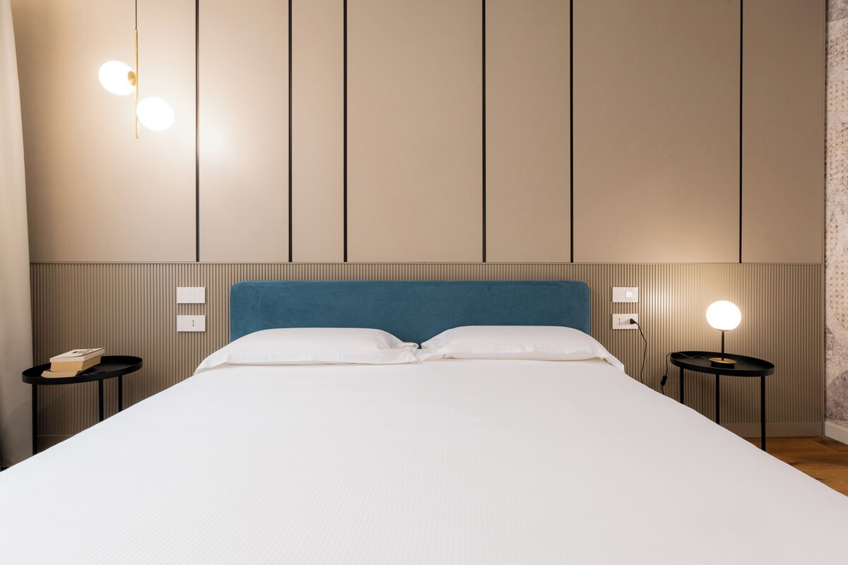 A hotel room with a large bed, two bedside tables, a blue headboard, and wall-mounted lighting.