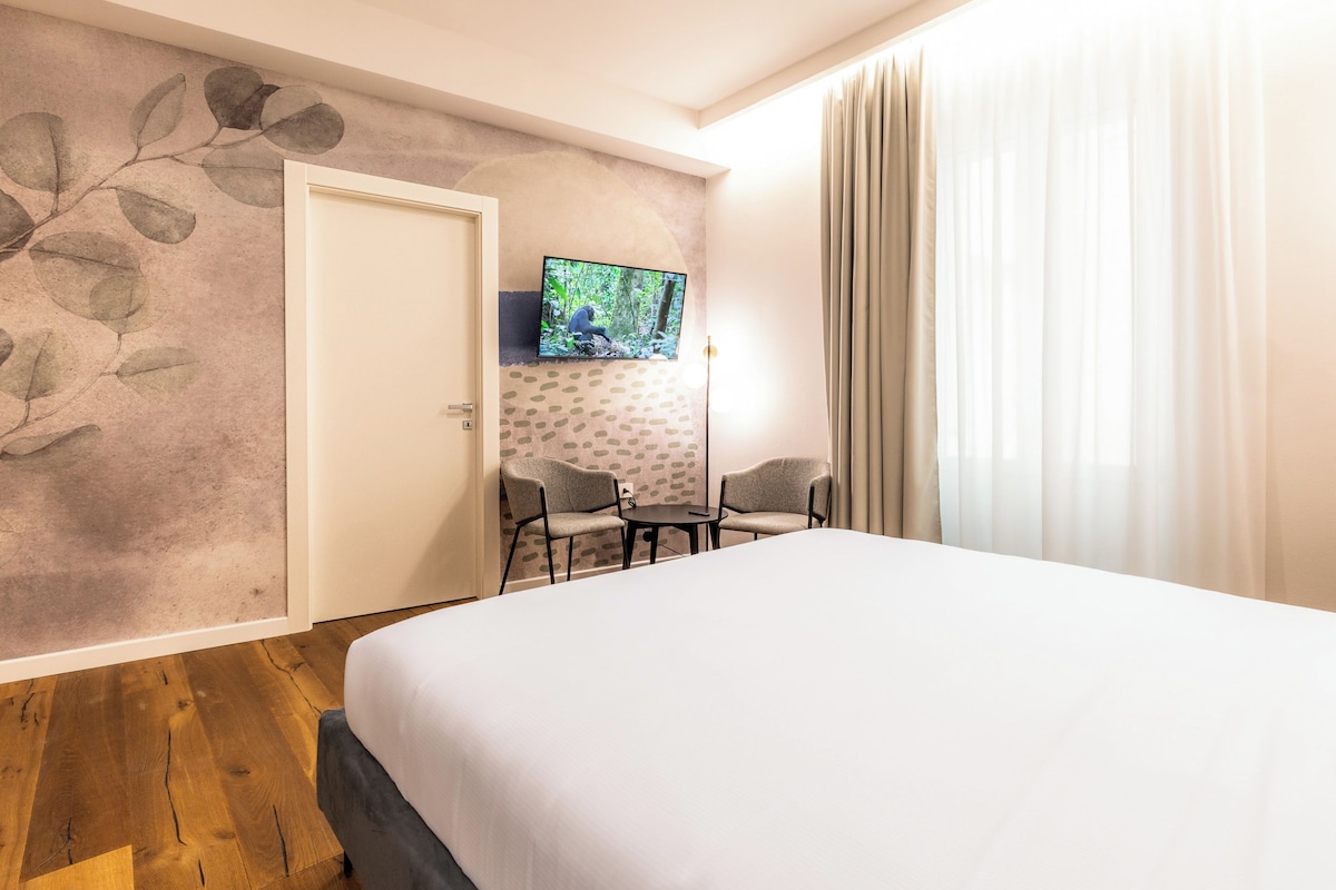 Una habitación de hotel con cama, televisor montado en la pared, dos sillas y una puerta.