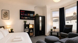 Ein Hotelzimmer mit einem Bett, einer Sitzecke mit Poufs, einer schwarzen Unterhaltungsecke inklusive Netflix-Anzeige, einem Spiegel und einem Fenster mit Vorhängen.