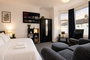 Habitación de hotel con cama, zona de estar con otomanas, mueble negro de entretenimiento con la función de Netflix, espejo y ventana con cortinas.