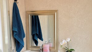 Casa de banho com lavatório branco, espelho com moldura dourada, toalha azul, dispensador de sabonete rosa e uma orquídea em vaso.