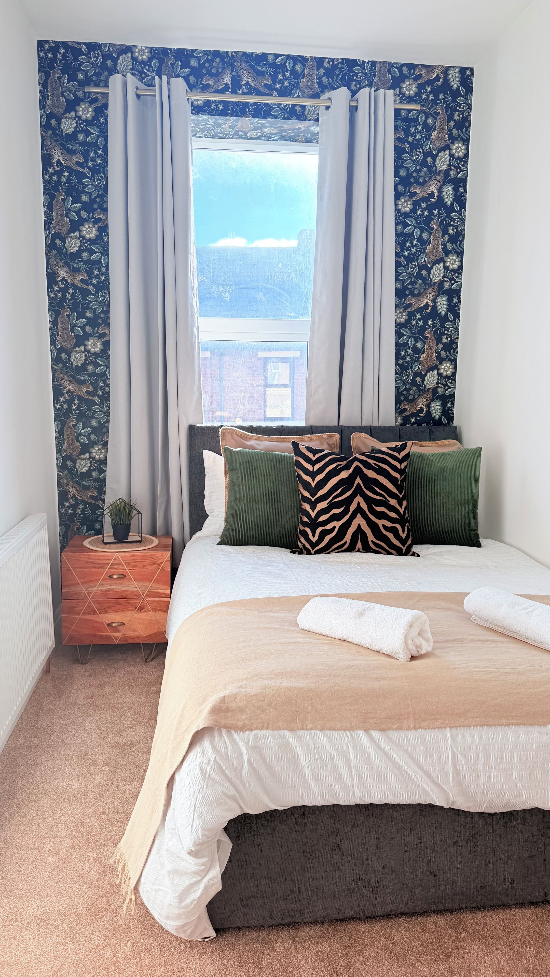 A bedroom with a bed, pillows, a nightstand, a window with curtains, and a view of the outside.