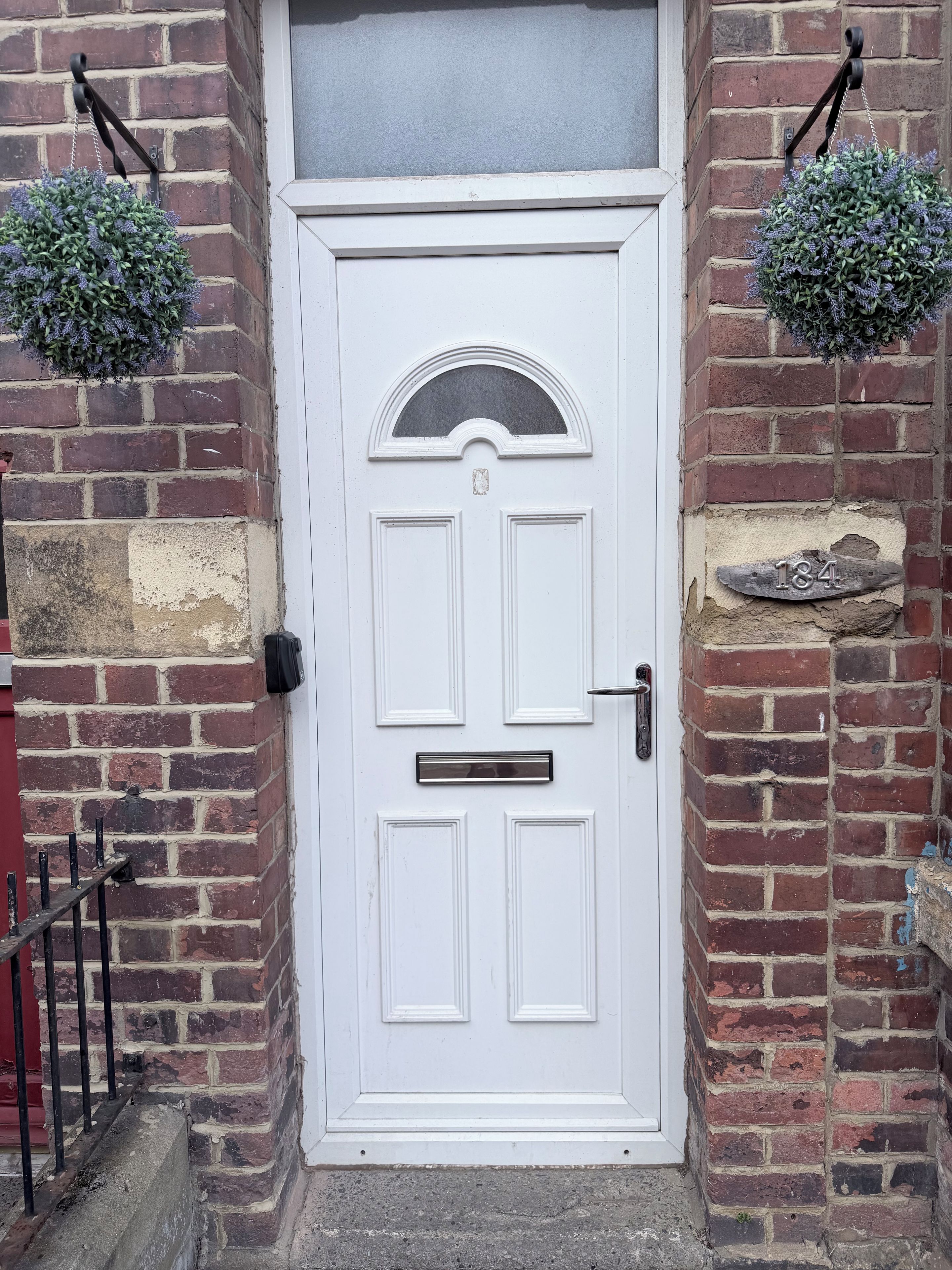 A white door with a mail slot and a number plaque, flanked by brick walls and hanging planters.