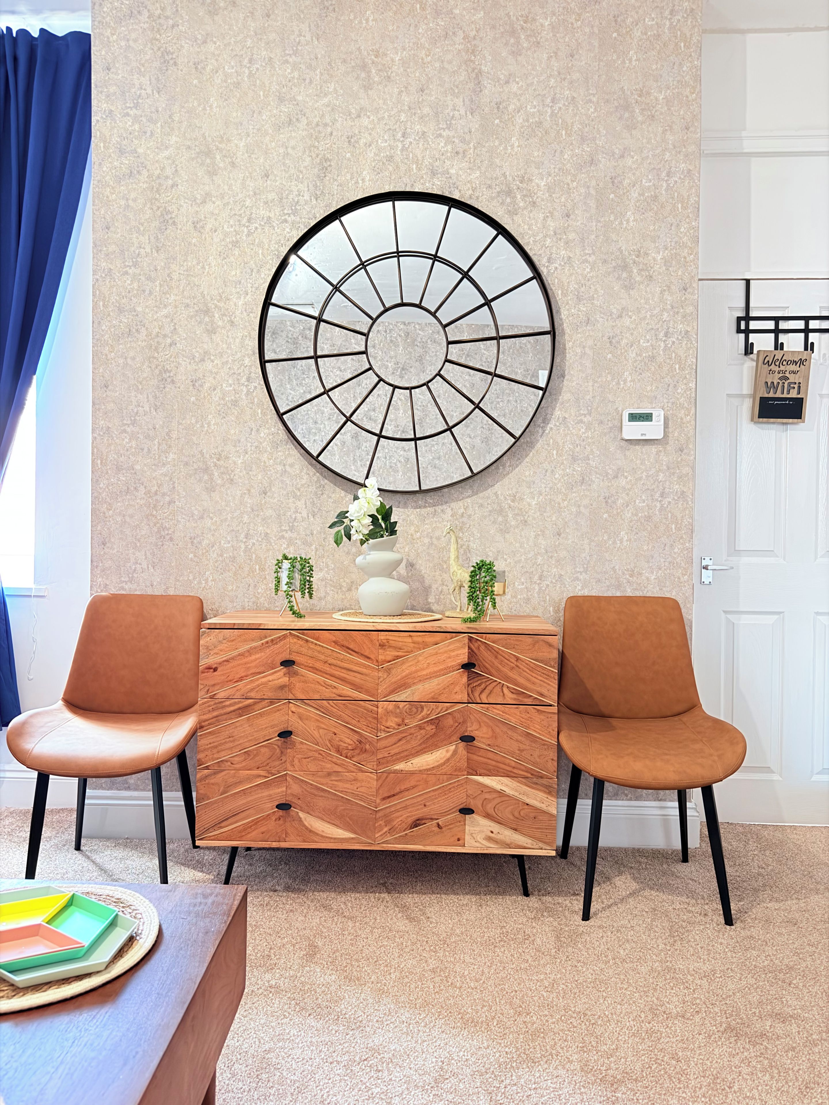 A modern room with a herringbone-patterned wooden dresser, two matching chairs, a large wall-mounted circular artwork, and a door with a welcome sign.