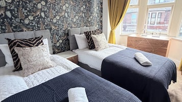 Hotellihuone, jossa on kaksi sänkyä, kuviollinen tapetti, ikkuna verhoilla ja näkymä ulkona oleviin rakennuksiin.