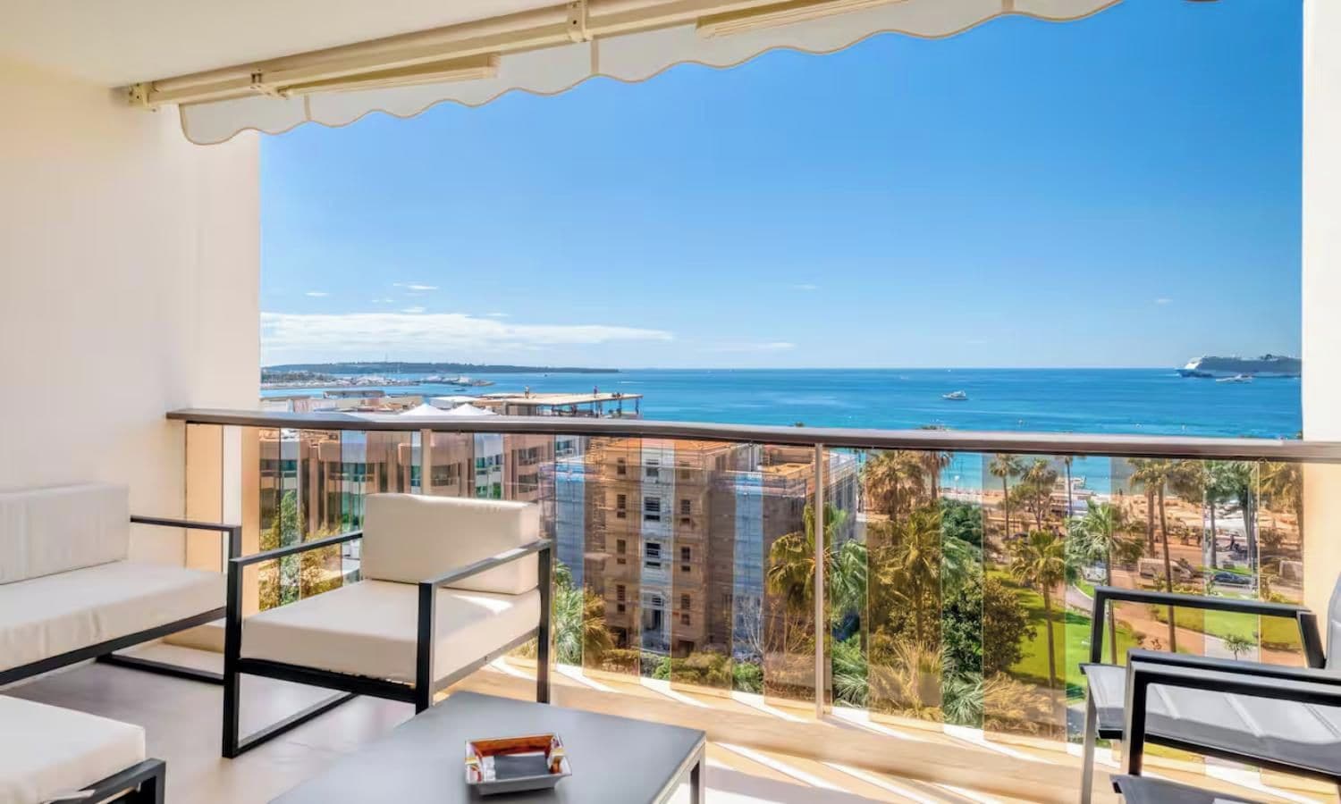 A balcony with a view of the sea and buildings. There are two white sofas and a small table with a tray.