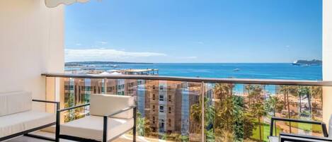 A balcony with a view of the sea and buildings. There are two white sofas and a small table with a tray.