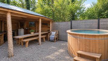 Un área al aire libre con jacuzzi de madera, una banca y una mesa, rodeada por una valla de madera y árboles.