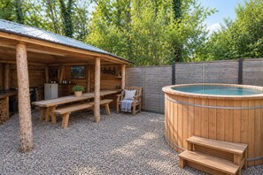 A wooden outdoor hot tub area with a wooden bench and a table, surrounded by a wooden fence and trees.
