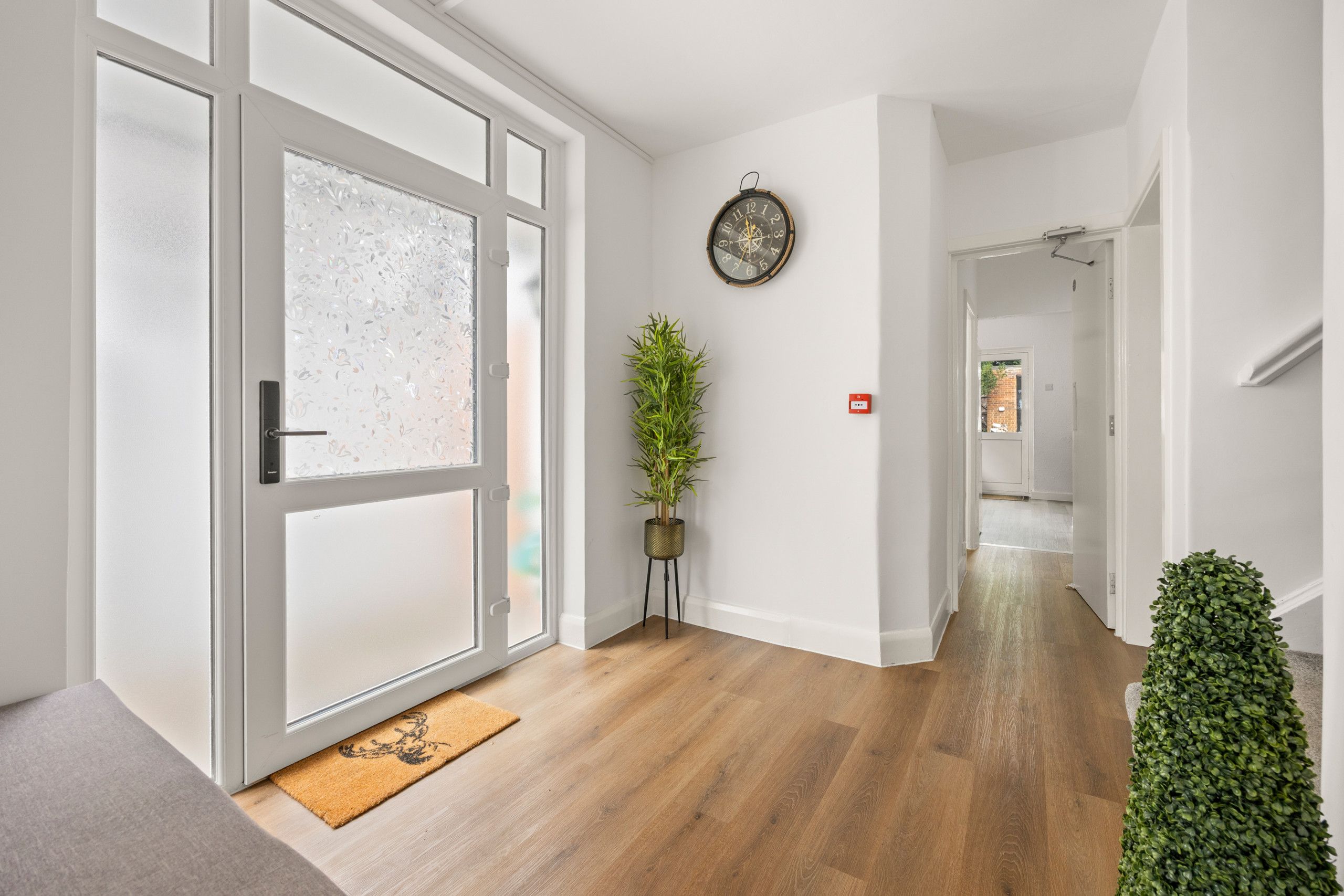 A hallway with a wooden floor, a door with frosted glass panels, a potted plant, and a wall-mounted clock.