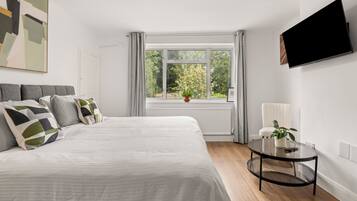 A bedroom with a bed, a TV mounted on the wall, a window with curtains, and a small table with a plant.