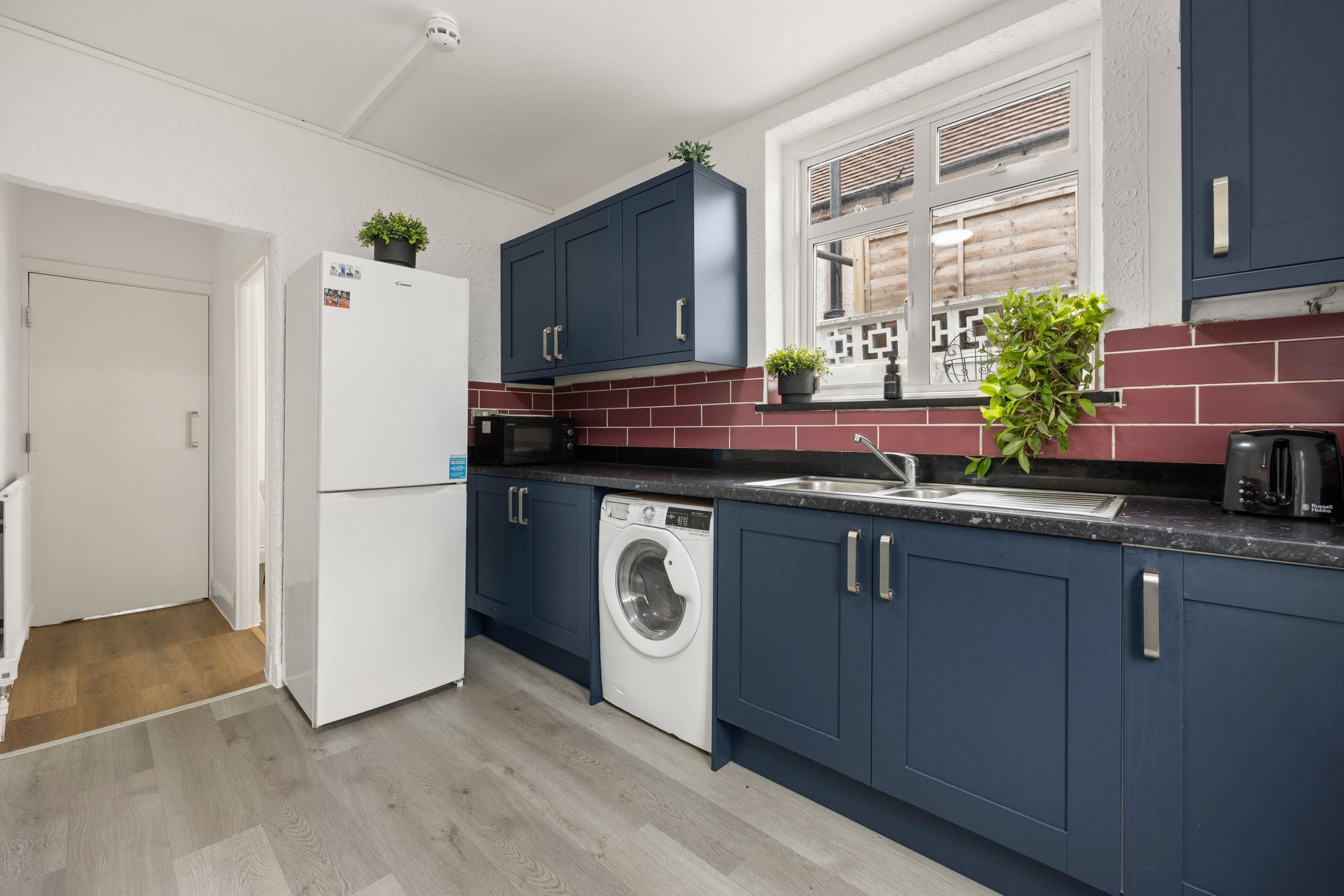 A modern kitchen with dark blue cabinets, a white refrigerator, a washing machine, and a window with a view of a wooden fence.