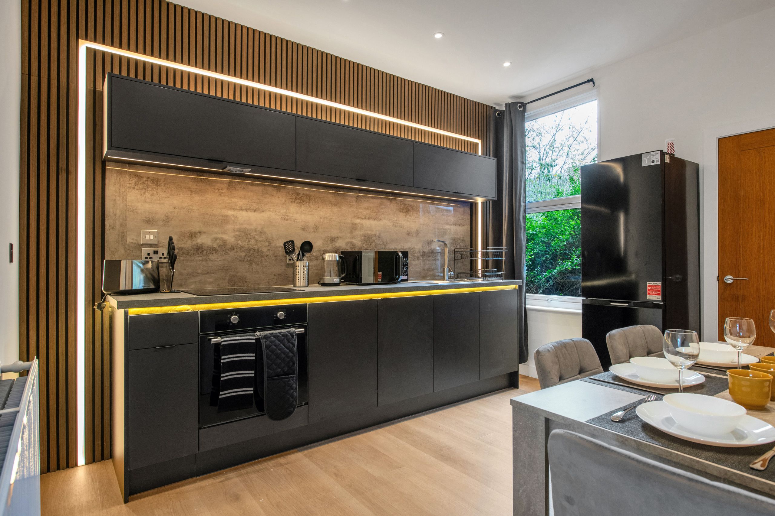 A modern kitchen with a central island, built-in appliances, and a dining area with a view of greenery.
