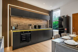 A modern kitchen with a central island, built-in appliances, and a dining area with a view of greenery. - K Suites - Modern Urban 3 bed House w/ Garden views (Nottingham)