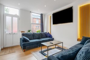 A modern living room with a sofa, a coffee table, and a TV mounted on the wall. - K Suites - Modern Urban 3 bed House w/ Garden views (Nottingham)