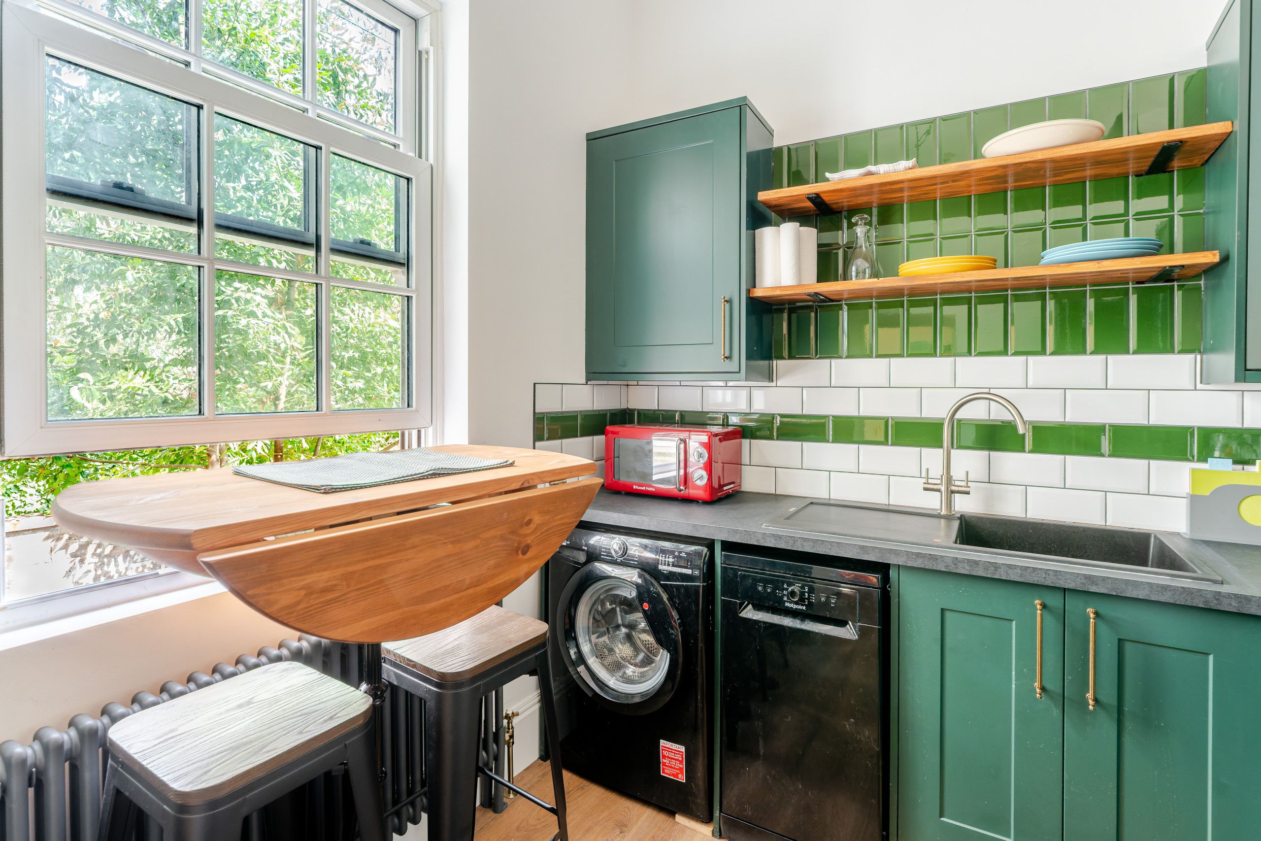 A compact kitchen with a built-in washing machine, a red toaster, and a window with a view of greenery.