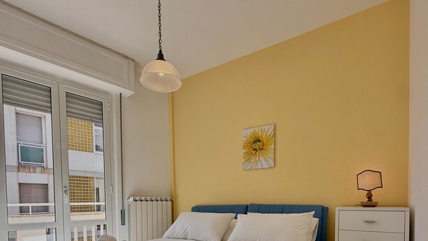A bedroom with a bed, a nightstand, a lamp, a window with blinds, and a radiator. - Apartment 'Casa Di Fiorentino' with Balcony, Wi-Fi and Air Conditioning (Sanremo)