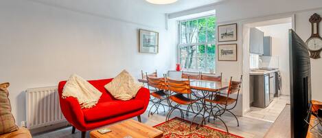 A living room with a red sofa, a wooden coffee table, and a dining area with a table and chairs. There is a large window with a view of trees and a red kettle on the windowsill.
