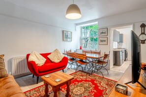 A living room with a red sofa, a wooden coffee table, and a dining area with a table and chairs. There is a large window with a view of trees and a red kettle on the windowsill. - K Suites - Chic 3 bed Apt w/ Garden views (Bristol)
