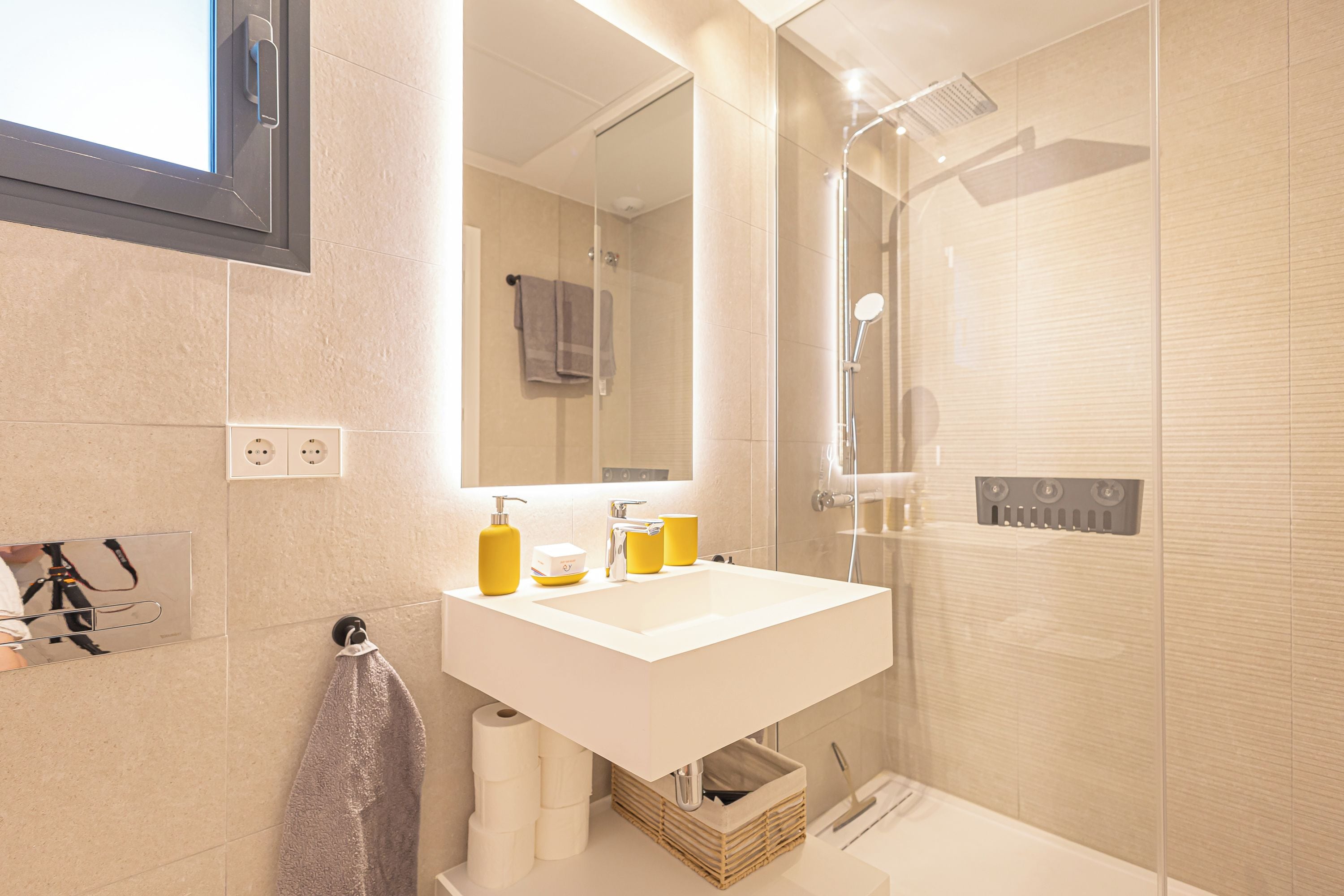 Un baño moderno con lavamanos, espejo y ducha blancos. Hay artículos de aseo en el lavamanos y un perchero para toallas.
